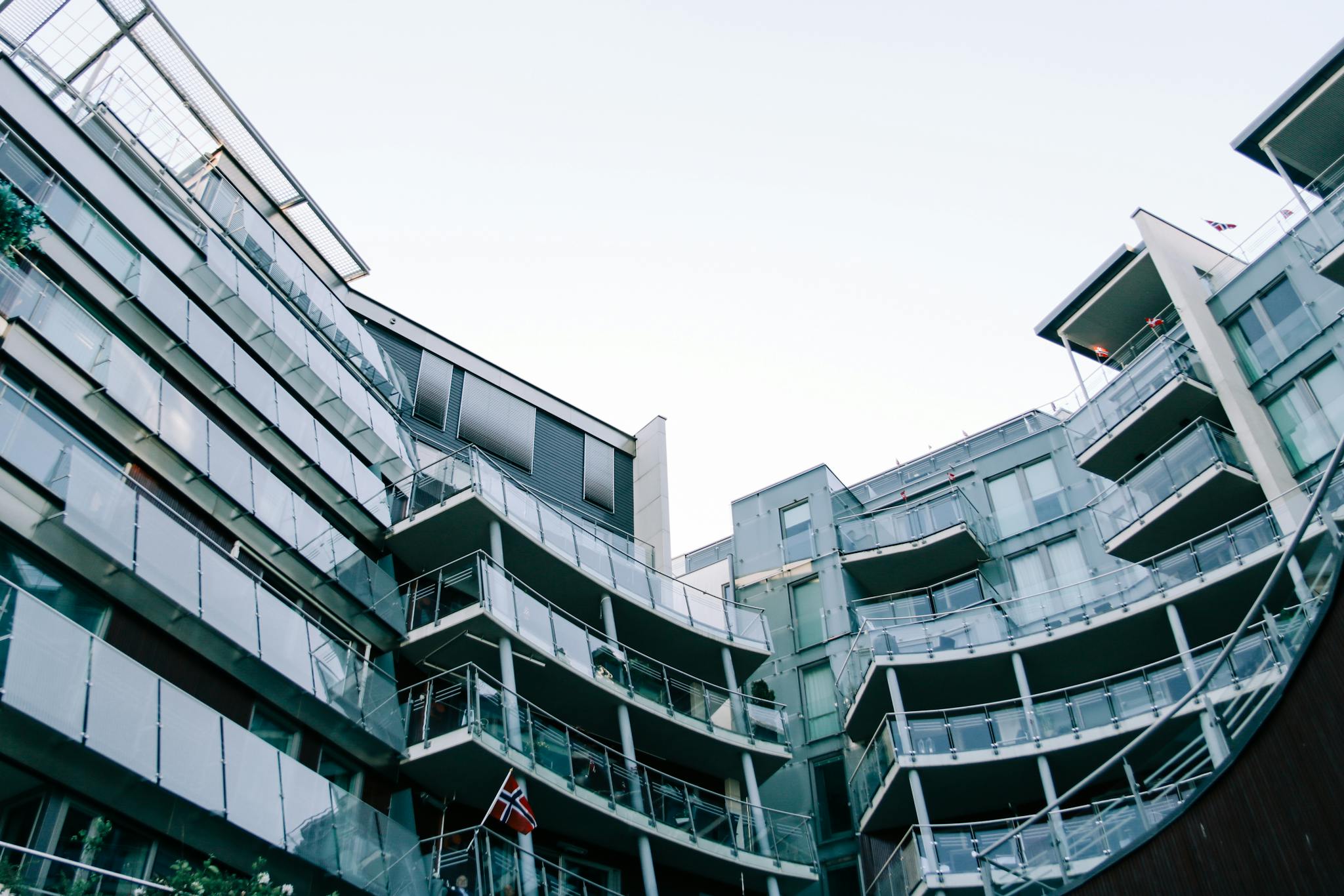 Low-angle view of a modern glass building featuring balconies and Norwegian flags.