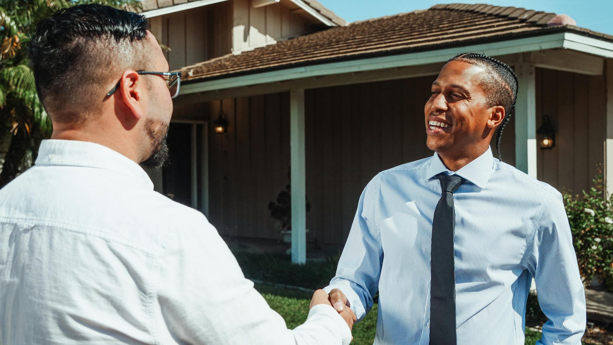 Two real estate professionals shaking hands in front of a new home during a transaction.